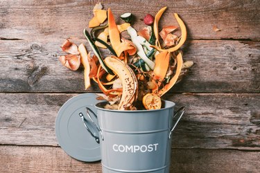 Peeled vegetables on chopping board, white compost bin on wooden background. Top view of kitchen food waste collected in recycling compost pot.