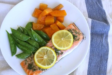 Overhead view of baked salmon topped with herbs and lemon slices, on a white plate with baked squash and snap peas