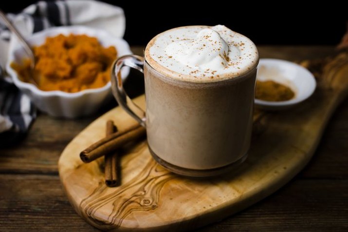 A pumpkin spice latte served in a glass mug on a cutting board, with spices and a dish of pumpkin puree visible in the background