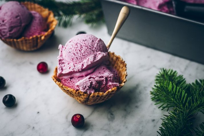 A waffle bowl of no-churn cranberry ice cream on a stone countertop, surrounded by loose berries, evergreen sprigs and more ice cream